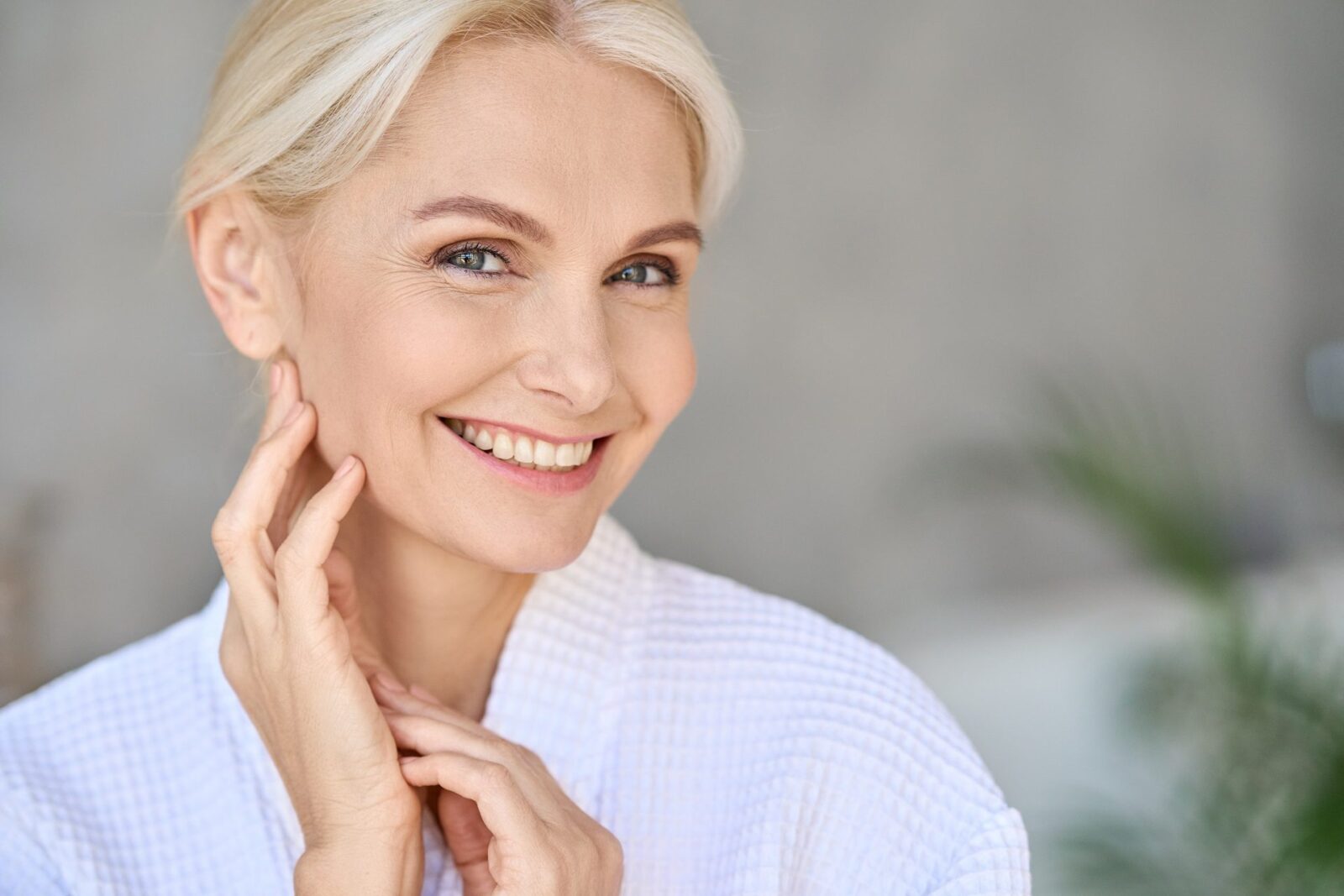 Closeup of happy smiling beautiful middle aged woman spa salon client wearing bathrobe looking at camera touching face. Spa procedures advertising. Skin care products concept. Copy space.