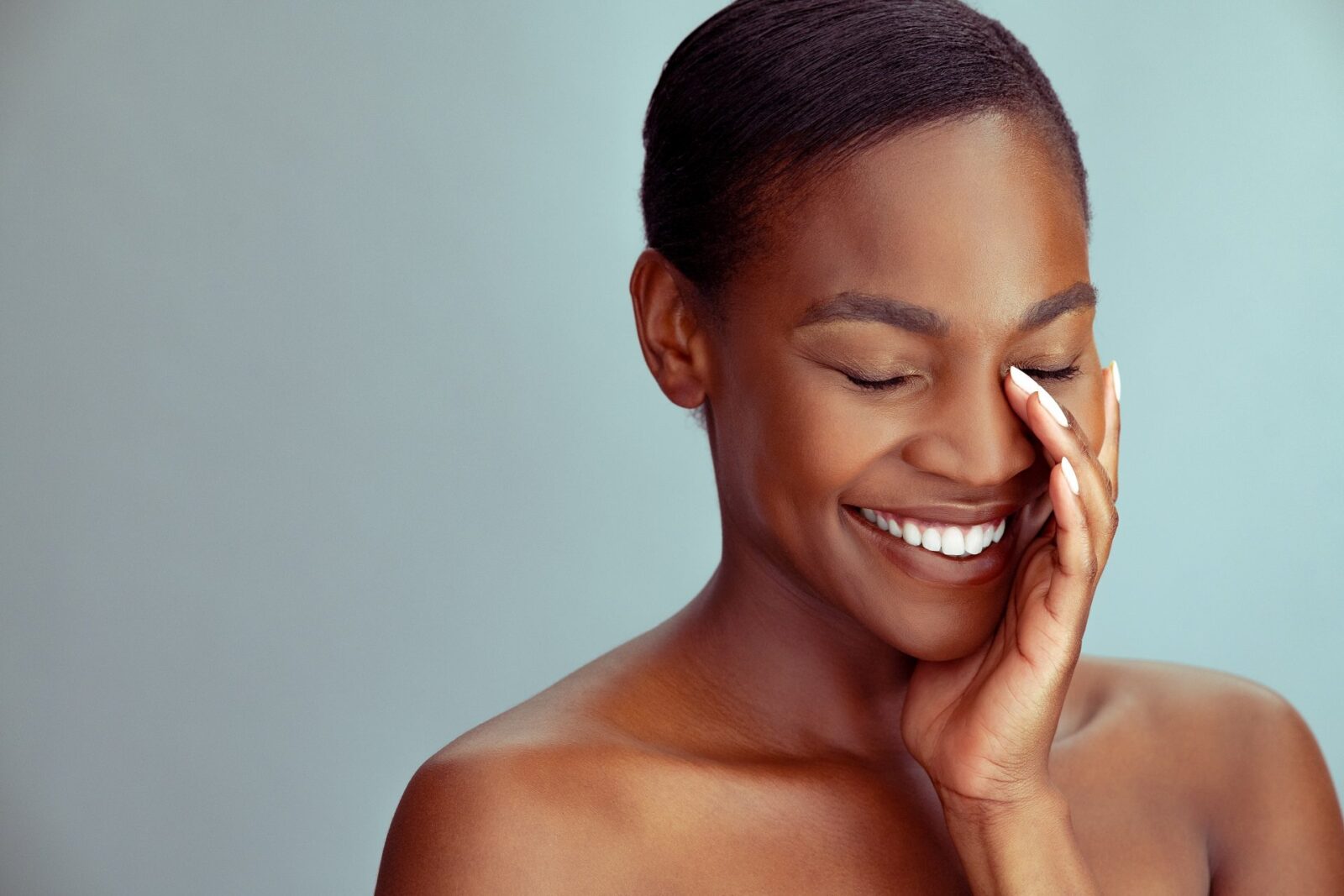 Smiling mature african woman with bare shoulder and eyes closed isolated against light blue wall. Black middle aged woman touching healthy skin against background with copy space. Beautiful mid african american lady with fresh glowing skin and bare shoulder looking down.