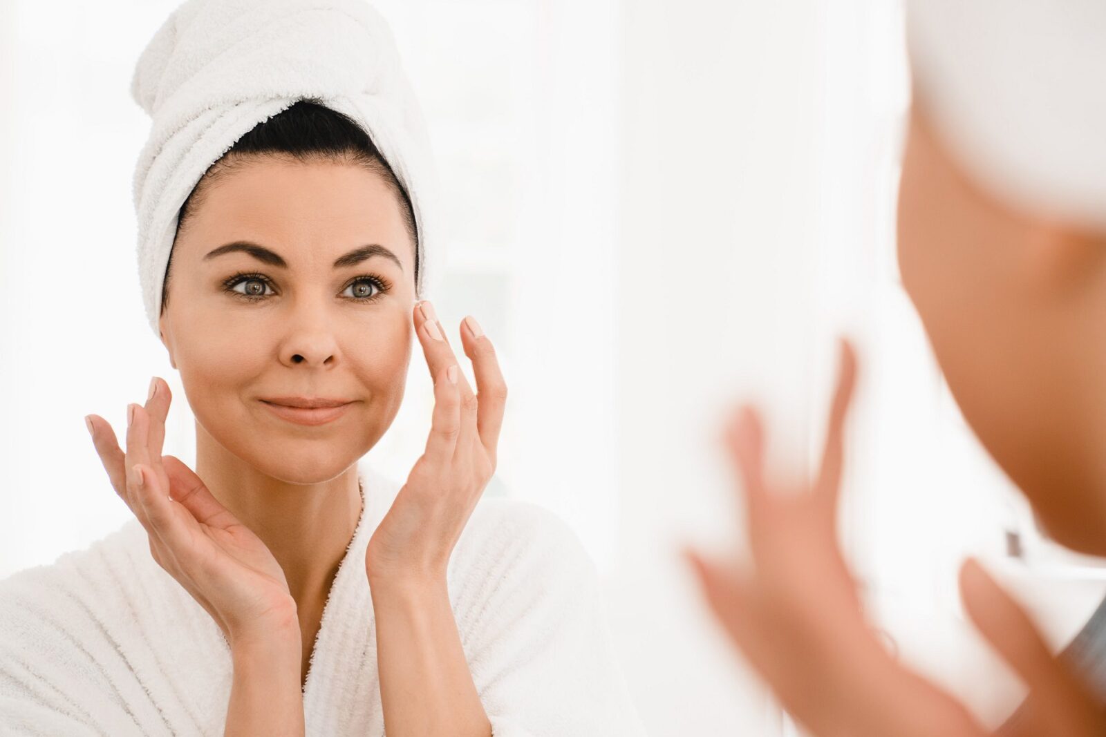 Good-looking caucasian middle-aged woman in turban and spa bathrobe after taking shower looking at the mirror while applying beauty creme moisturizer for anti-age anti-wrinkle effect